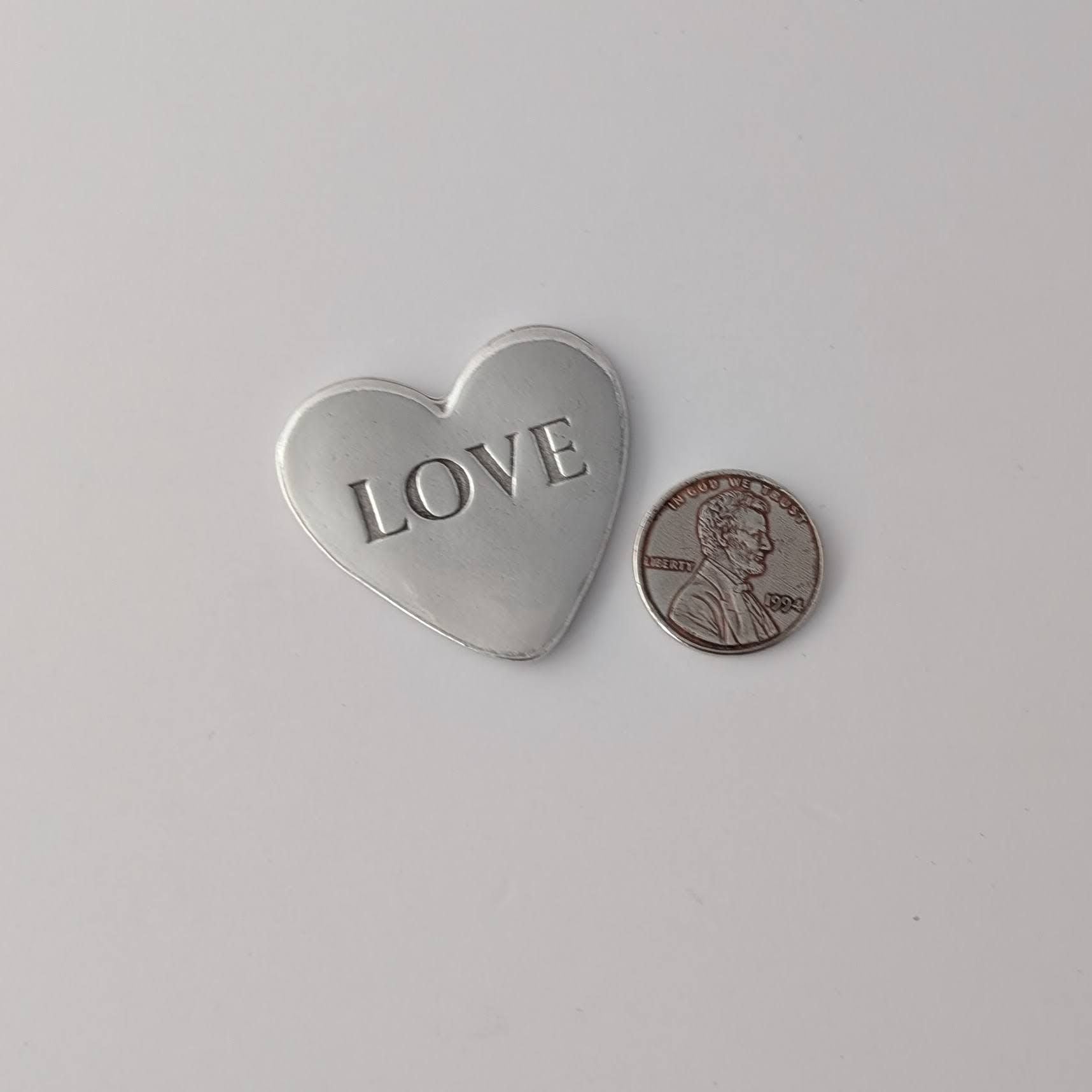 Silver heart-shaped pendant with 'LOVE' engraving next to a penny on a white background