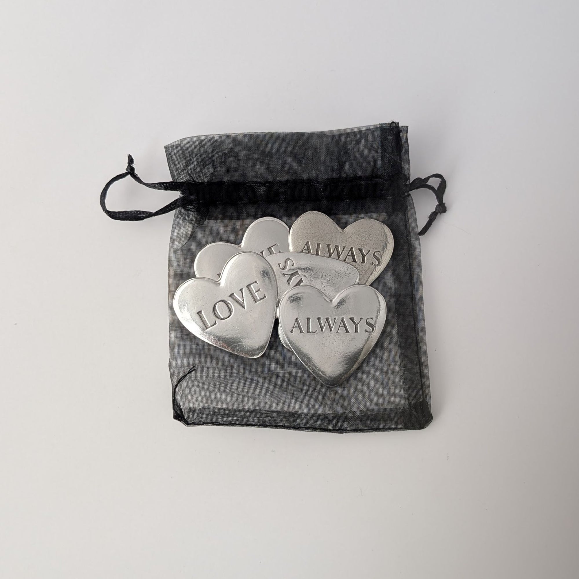 Silver heart-shaped tokens with engraved text on a black drawstring bag against a light gray background