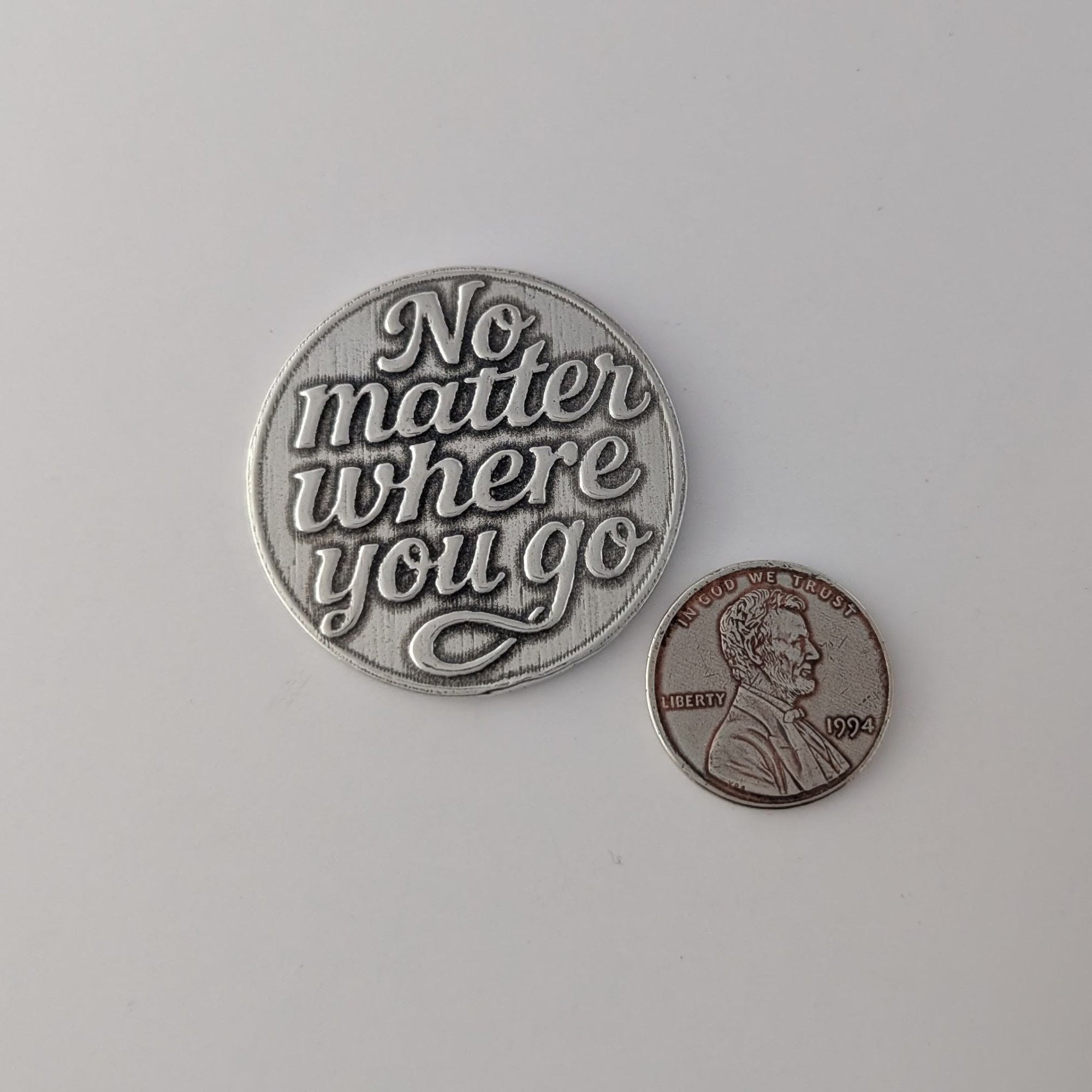 Pewter coin with 'No matter where you go' text next to a penny on a light gray background