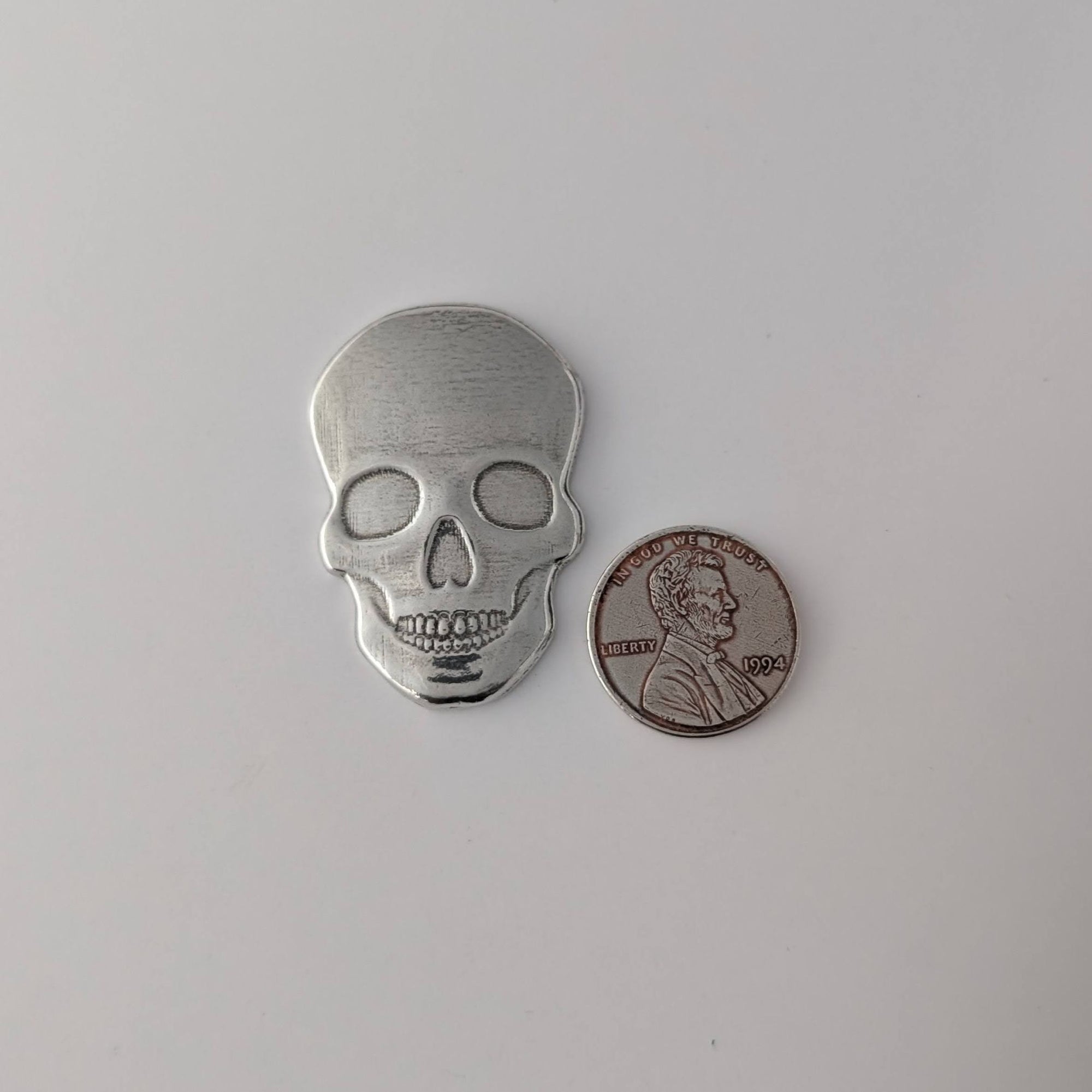 Silver skull-shaped object next to a penny on a light gray background