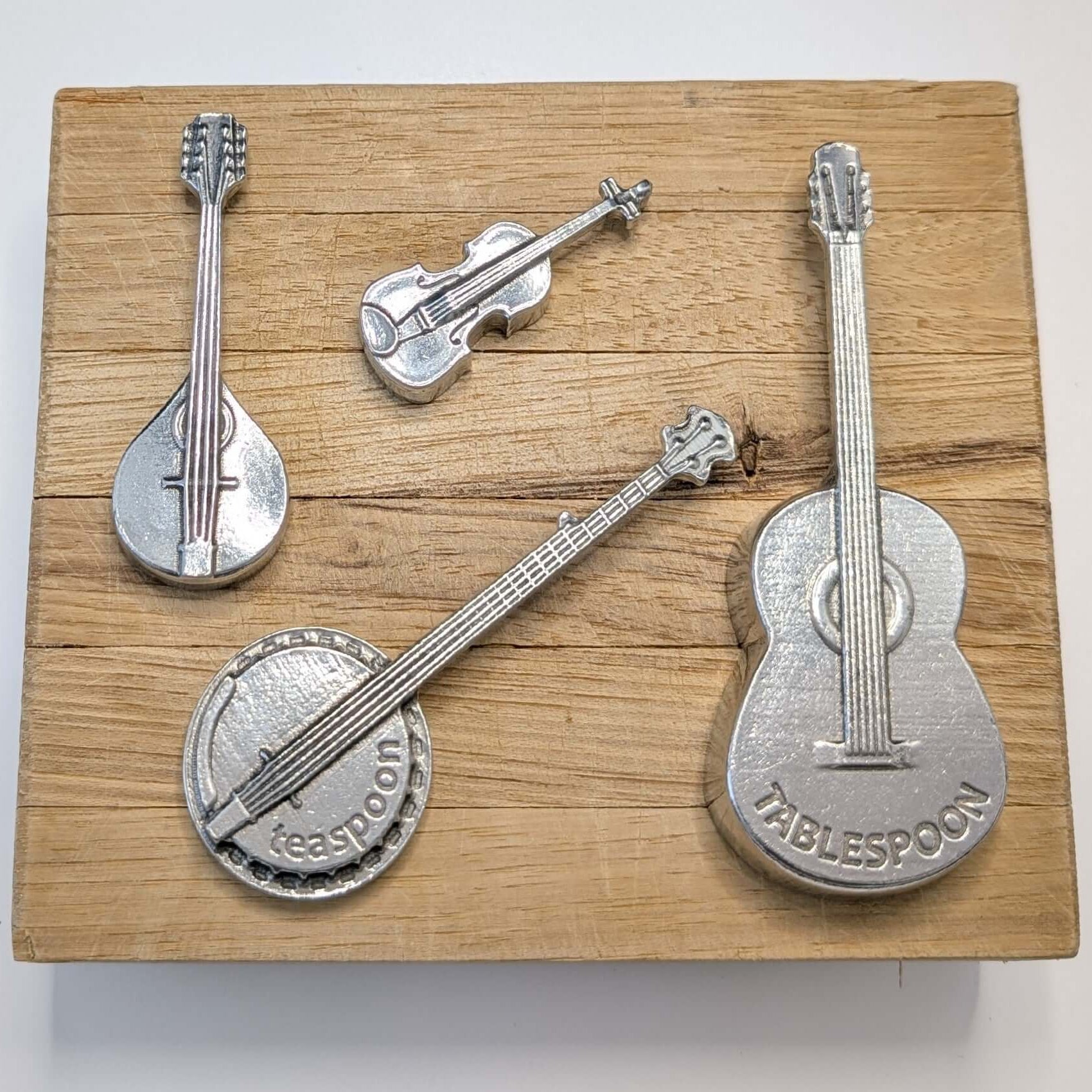 A set of four pewter measuring spoons designed to look like musical instruments, including a guitar, banjo, mandolin, and fiddle.
