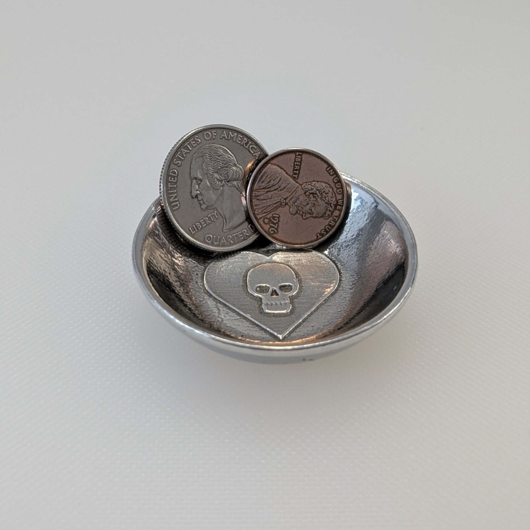 Skull in heart pewter ring dish filled with a quarter and penny on white surface.
