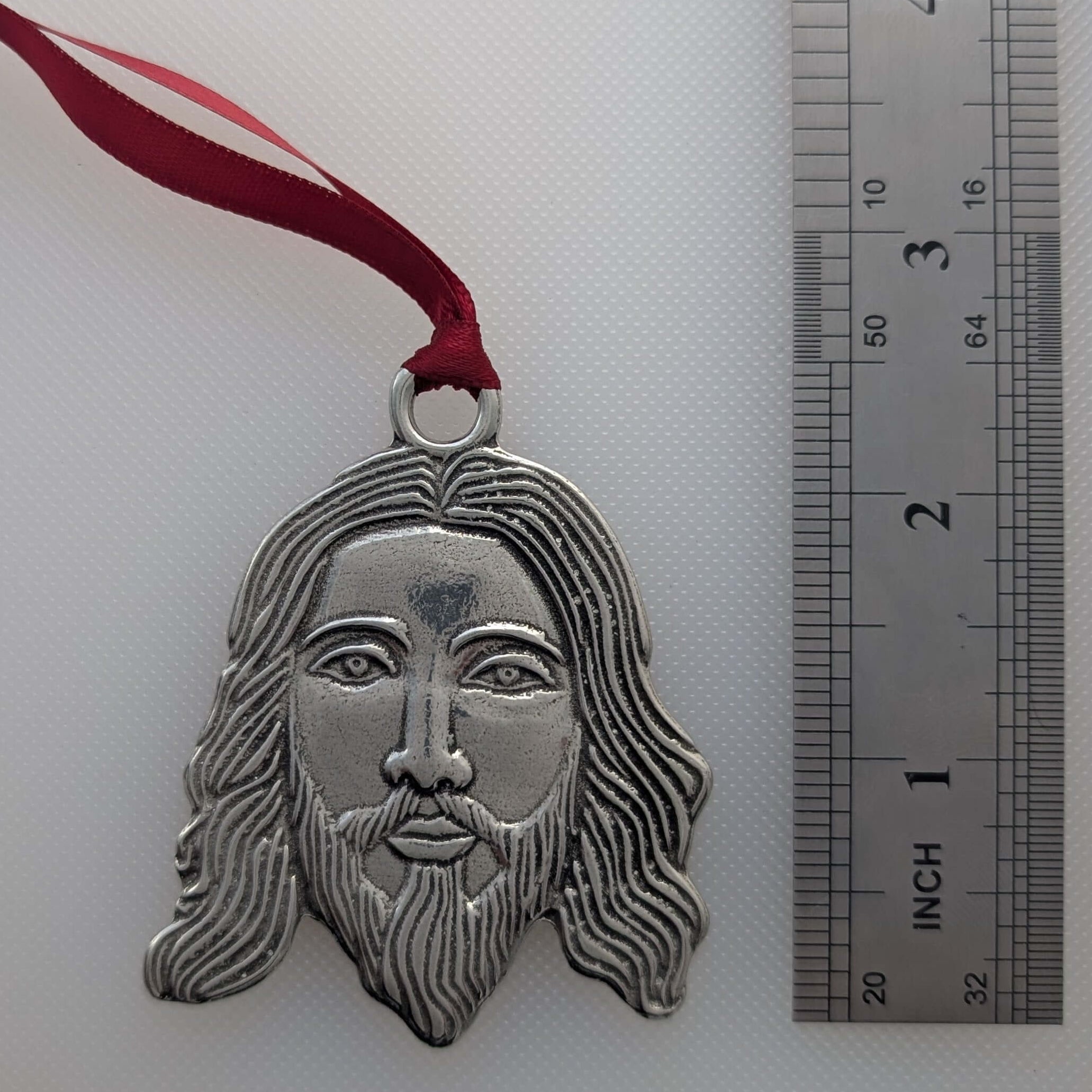 Silver pendant of a face with long hair and beard on a white background with a ruler.