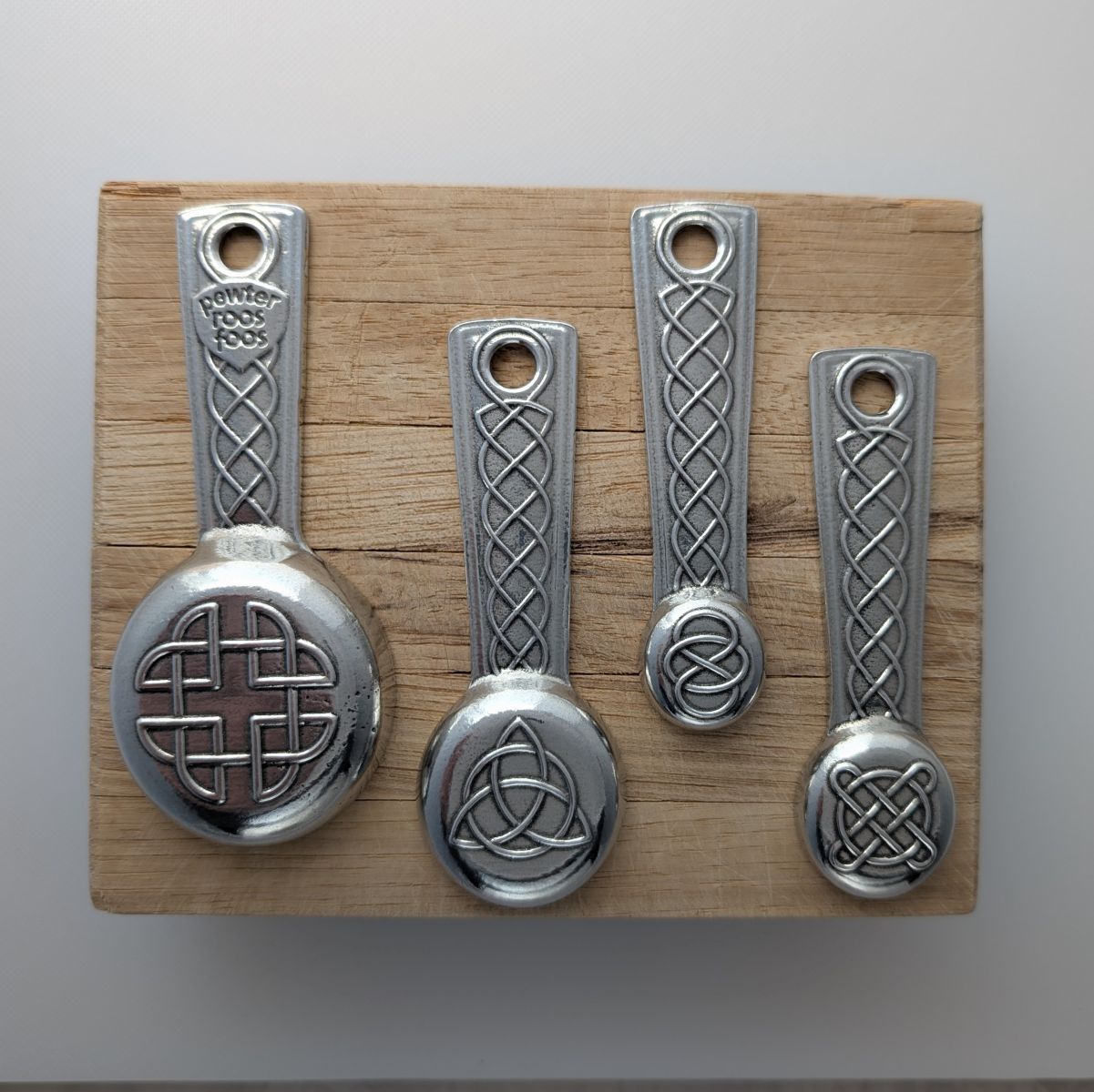 Set of four pewter measuring spoons with Celtic knot designs on a wooden board.
