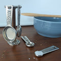 Set of silver measuring spoons on a wooden surface with a blue bowl and wooden spoon in the background.