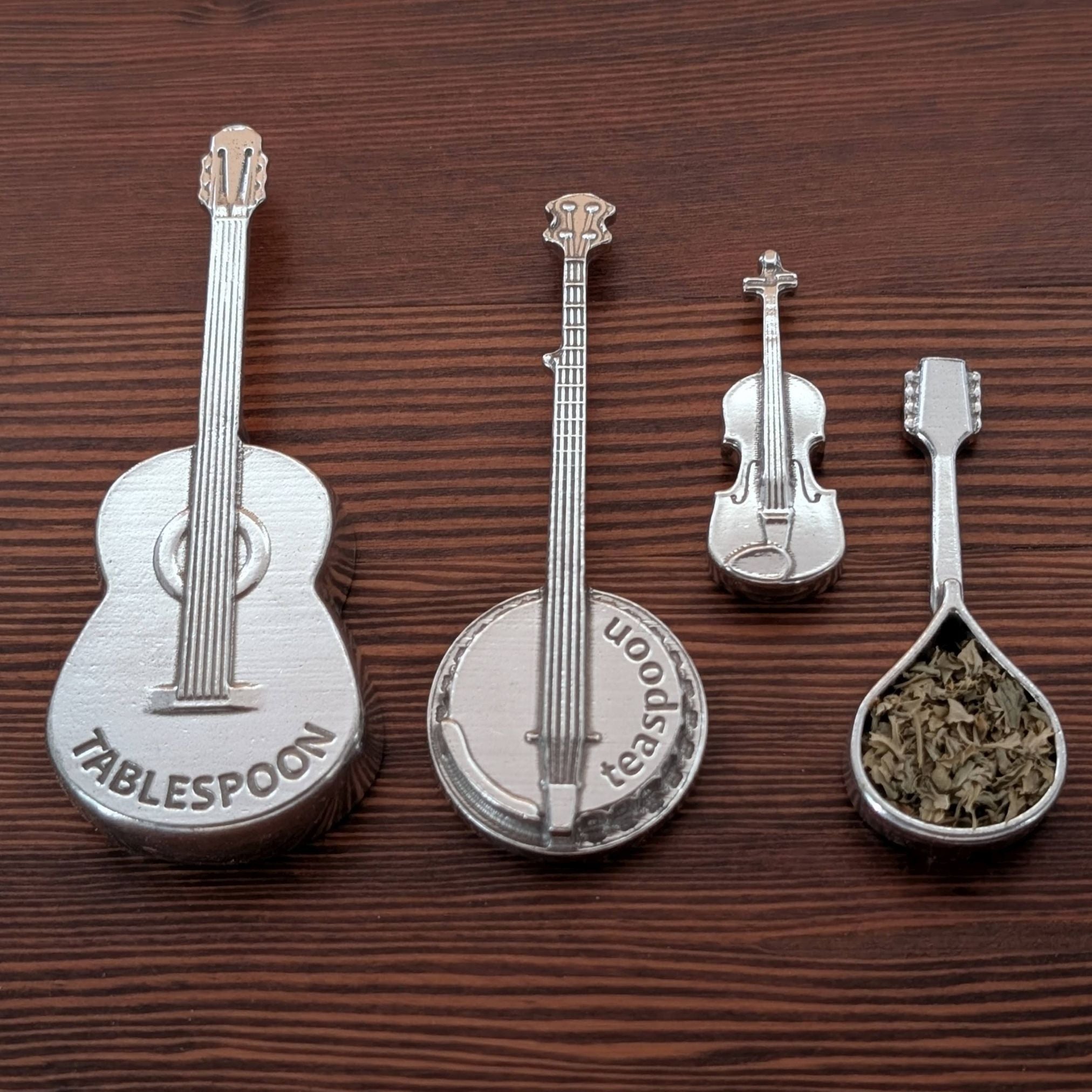 Metallic kitchen spoons shaped like a guitar, banjo, violin, and spoon on a wooden surface.