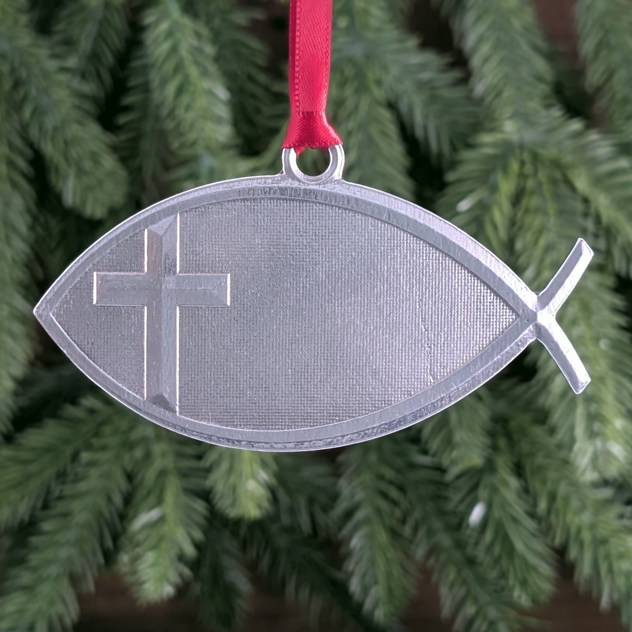 Silver fish-shaped ornament with a cross on a red ribbon against a green Christmas tree background