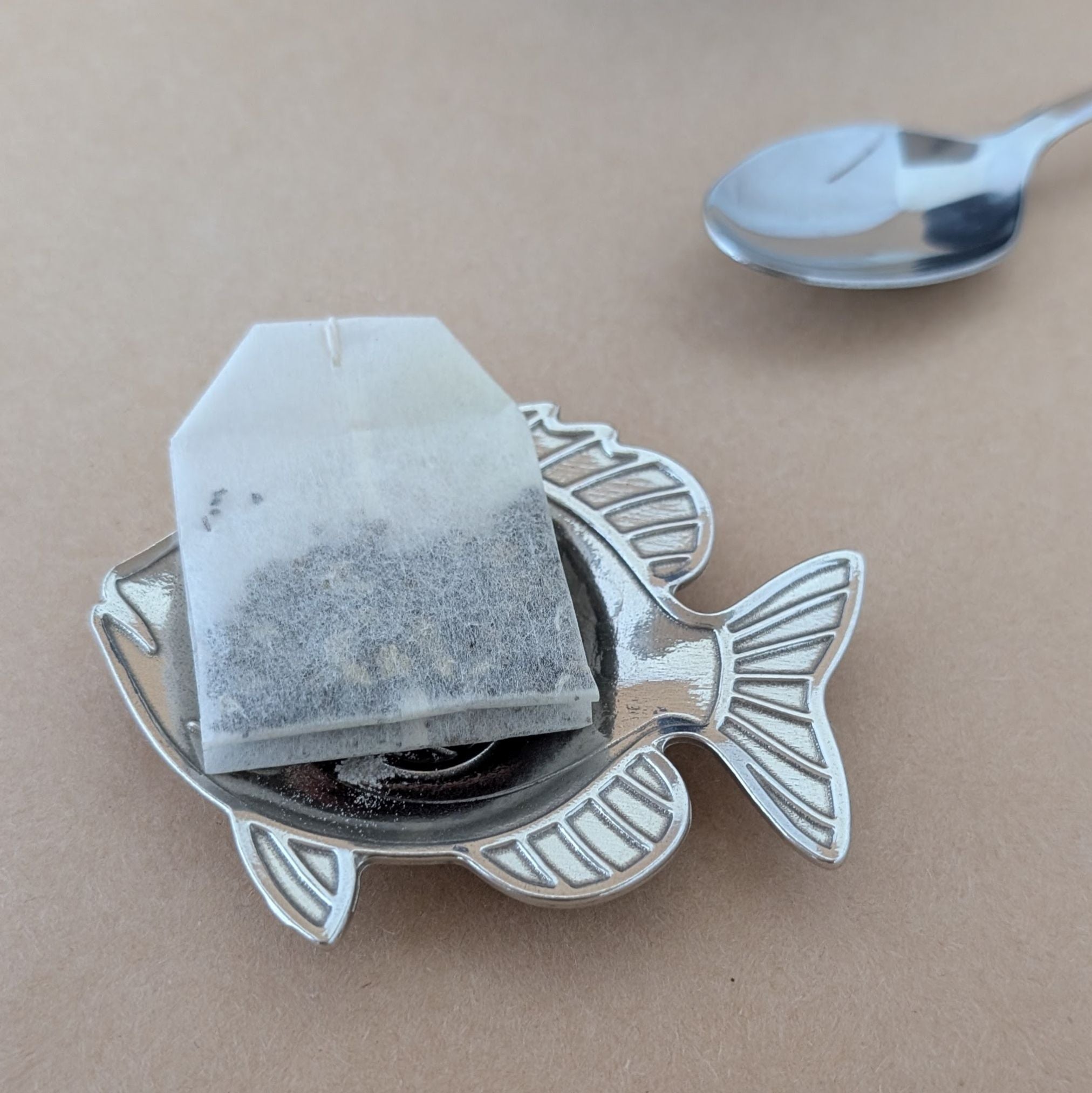 Tea bag in a fish-shaped teabag holder on a beige surface with a spoon in the background.