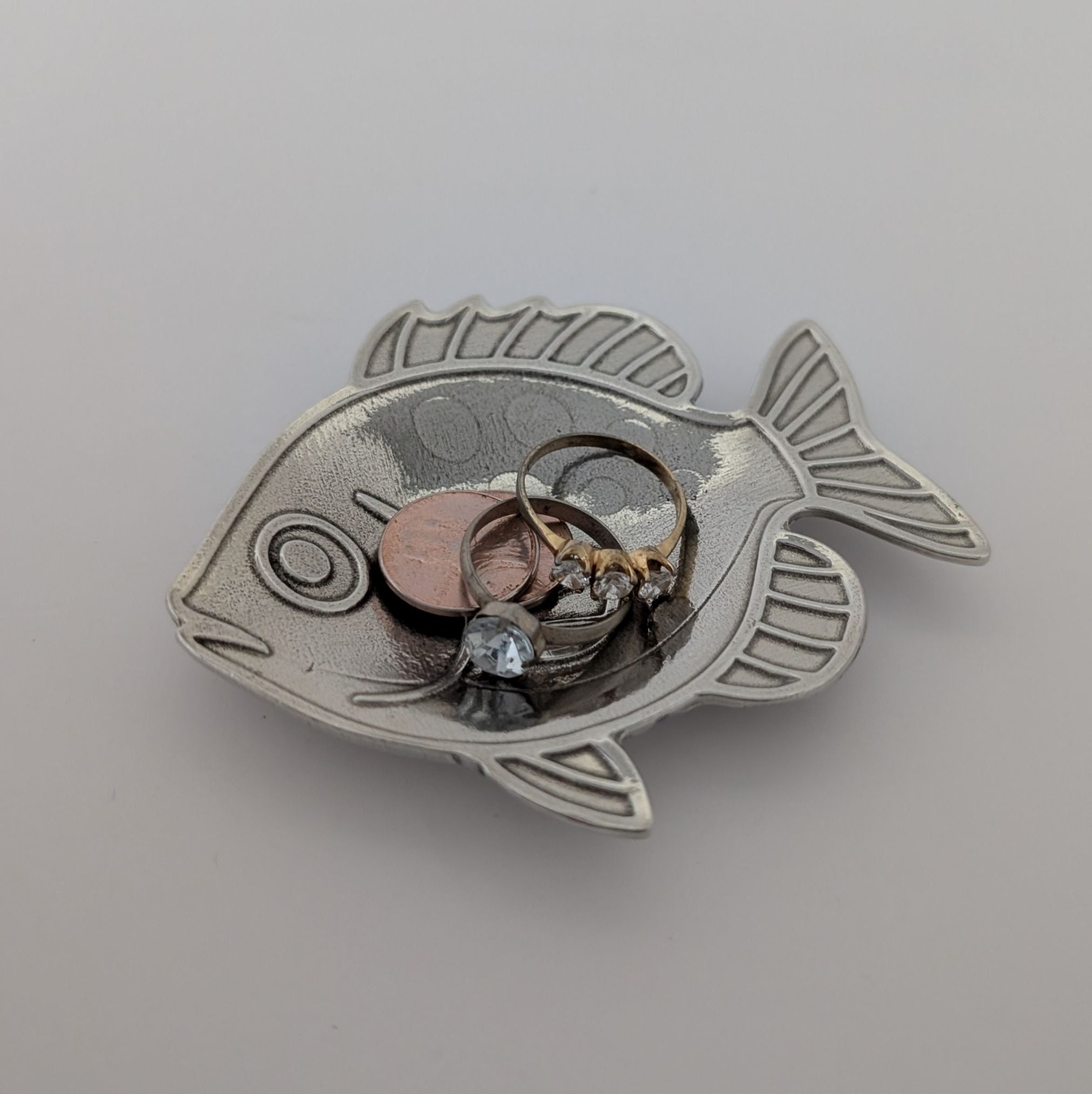 Silver fish-shaped jewelry holder with small items inside on a light gray background