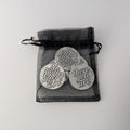 Three pewter circular charms with engraved text in a black drawstring bag on a light gray background