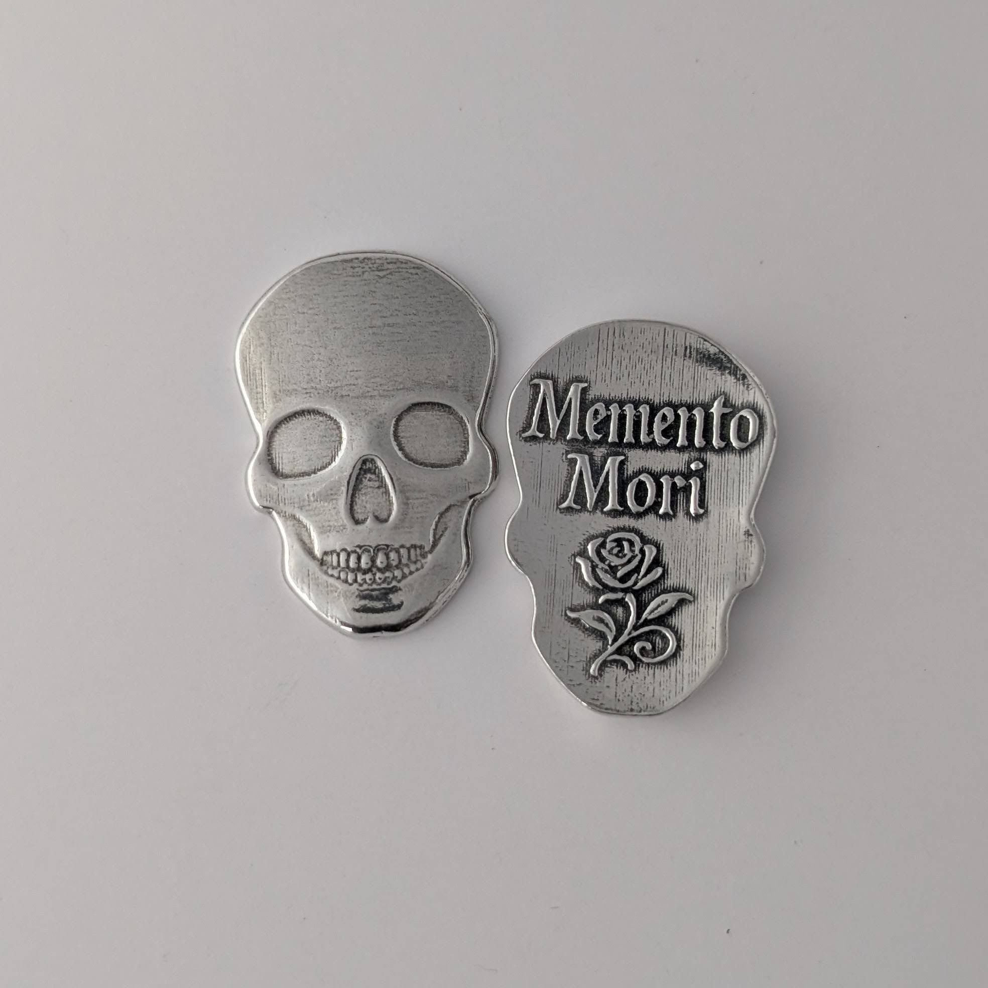 Silver skull-shaped pendant with 'Memento Mori' text and rose design on a gray background