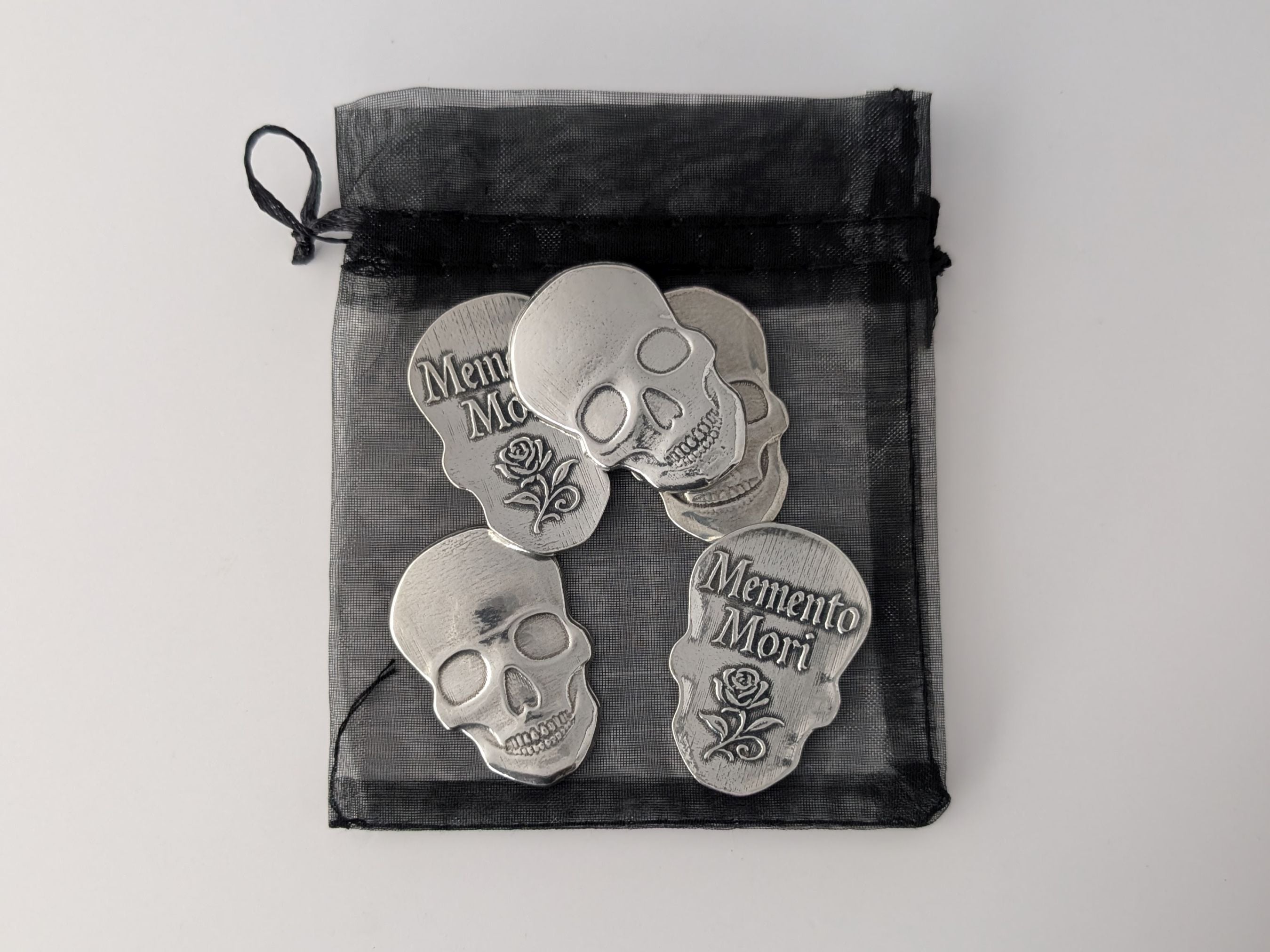 Silver skull-shaped 'Memento Mori' tokens in a black drawstring bag on a light gray background