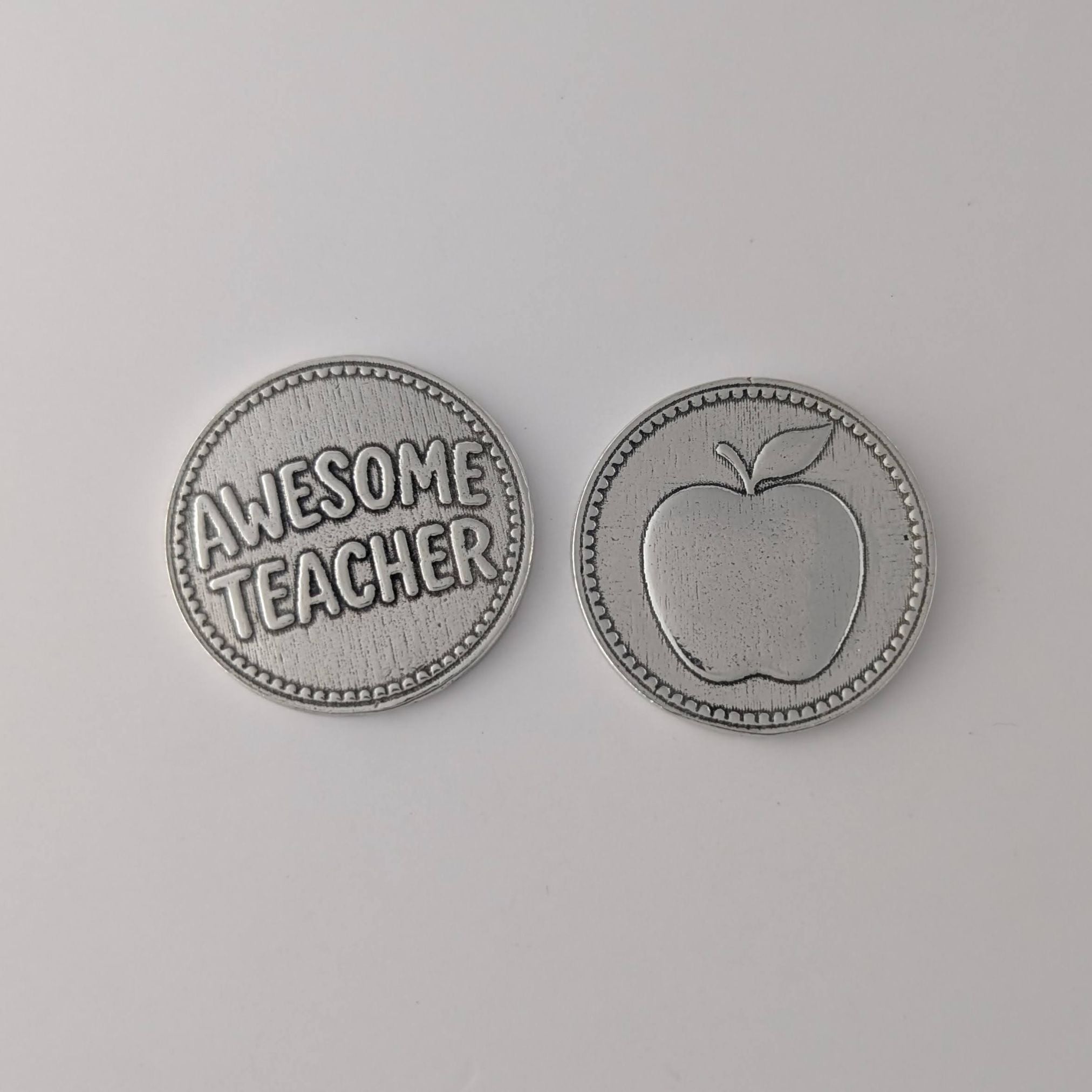 Two pewter coins on a gray background, one with 'Awesome Teacher' text and the other with an apple design.
