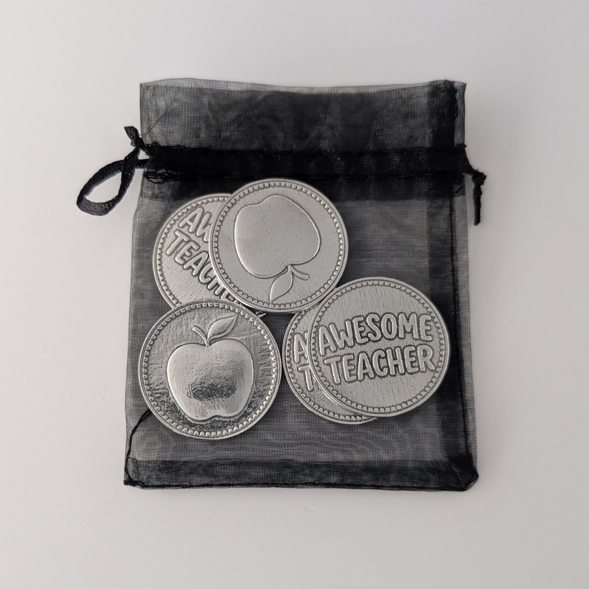 Four 'Awesome Teacher' coins with apple designs in a black drawstring bag on a light gray background