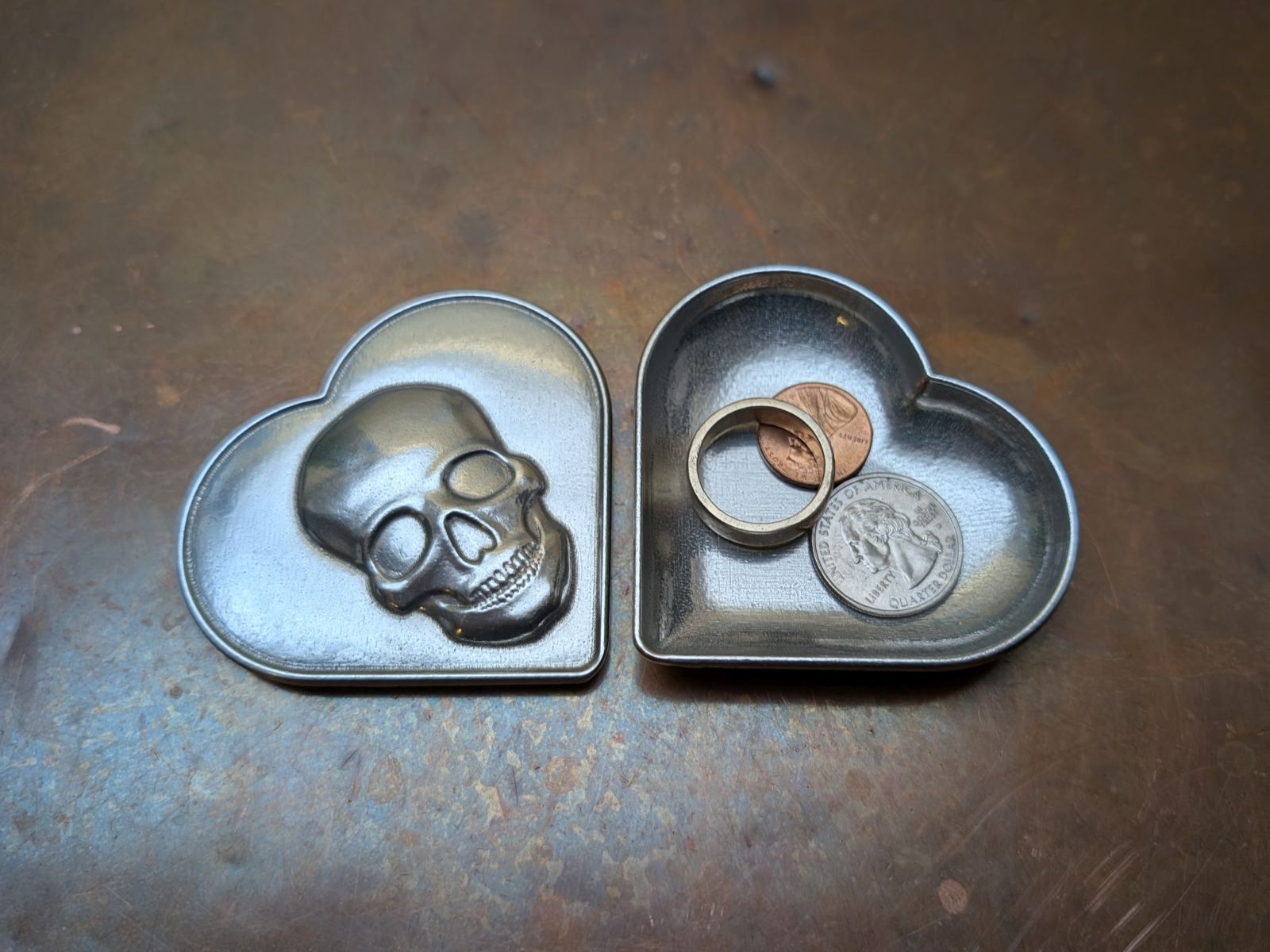Heart-shaped pewter container with skull design and coins on a copper surface
