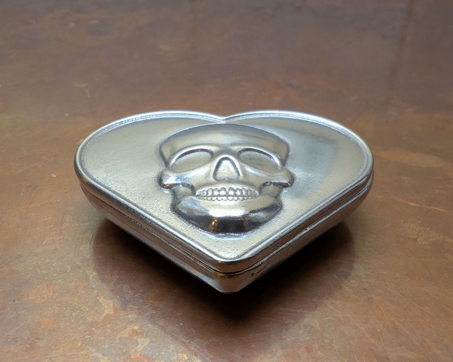 Heart-shaped pewter box with skull design on a brown surface