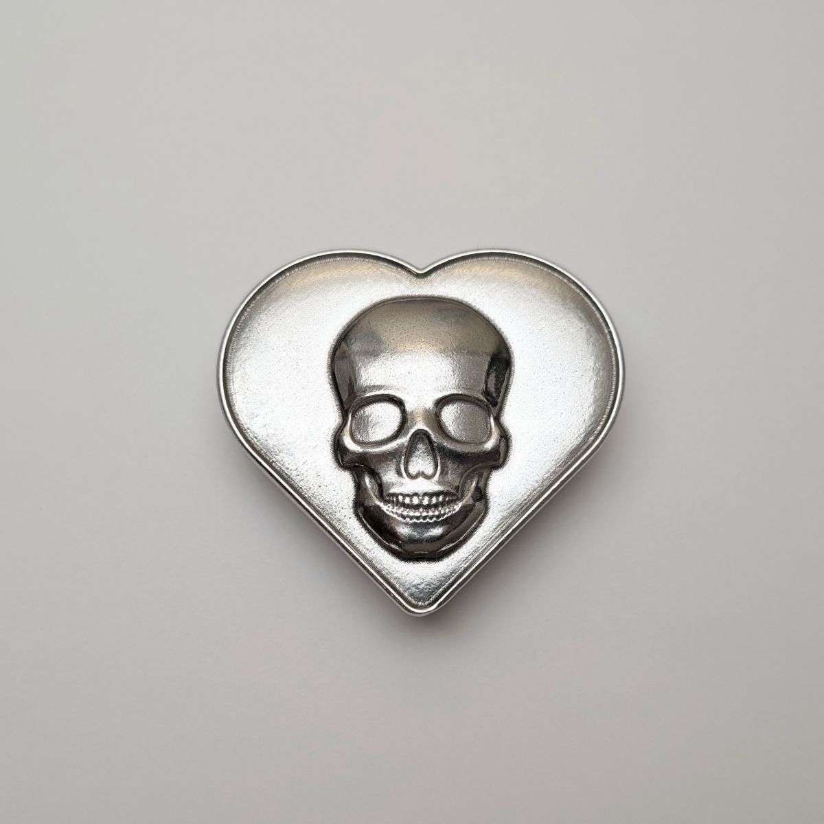 Silver skull design on a heart-shaped pewter box against a gray background