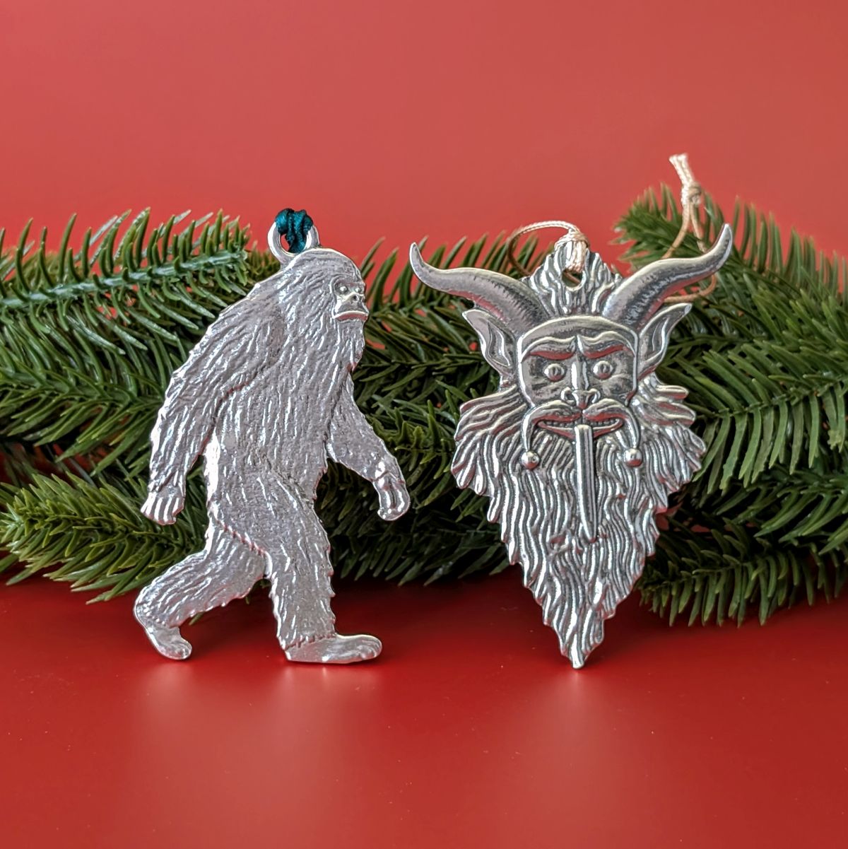 Two Christmas ornaments, one resembling a bigfoot and the other a horned figure, on a red background with greenery.