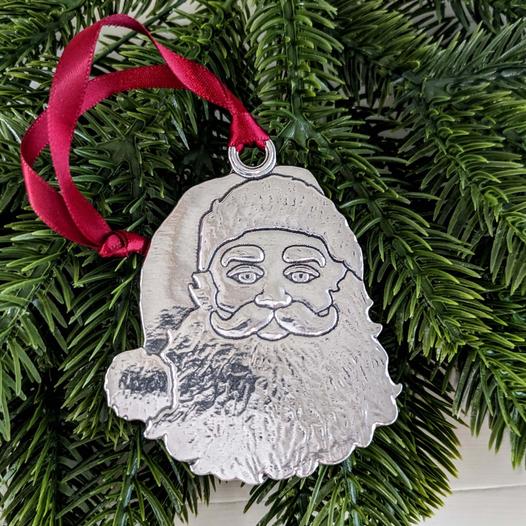 A handcrafted pewter Santa Claus ornament with a red hanging ribbon, displayed against a greenery background.