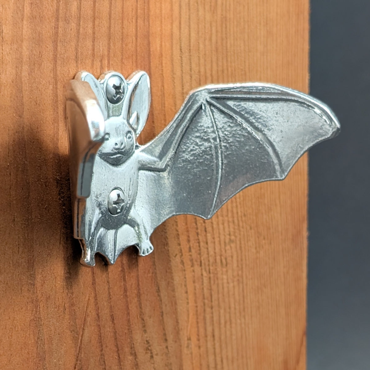 Pewter bat wall hook mounted on a wood board, angled close-up showing the wings and mounting screws